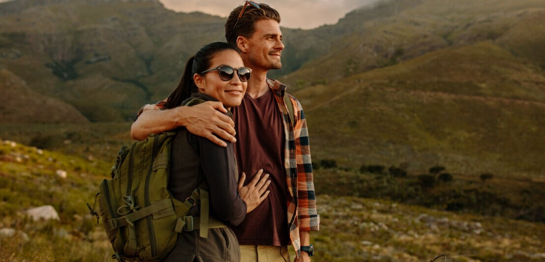 Ein Paar mittleren Alters in Freizeitkleidung umarmt sich während einer Wanderung und schaut zufrieden in die Berge 