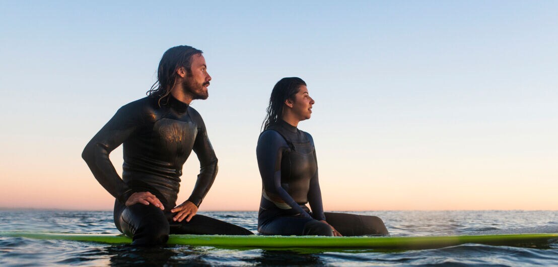 Ein junges Paar in Surfanzügen sitzt auf seinen Surfbrettern auf dem stillen Meer und blickt in die Ferne