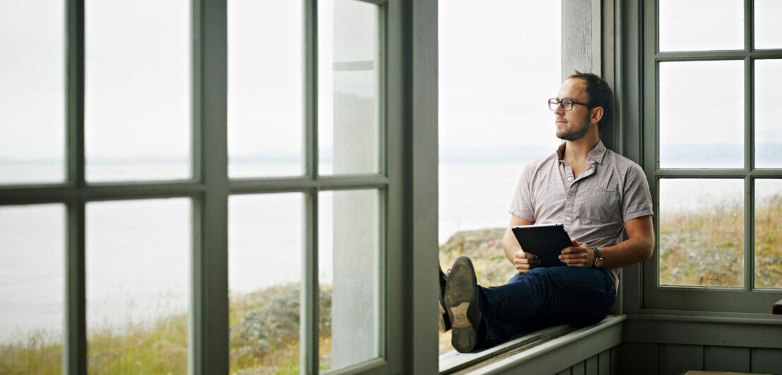Ein Mann sitzt mit einem Tablet auf einer Fensterbank und blickt aufs Meer hinaus