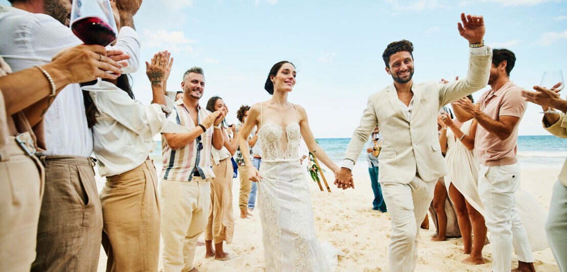 Ein Brautpaar rennt am Strand zwischen den klatschenden Gästen auf die Kamera zu