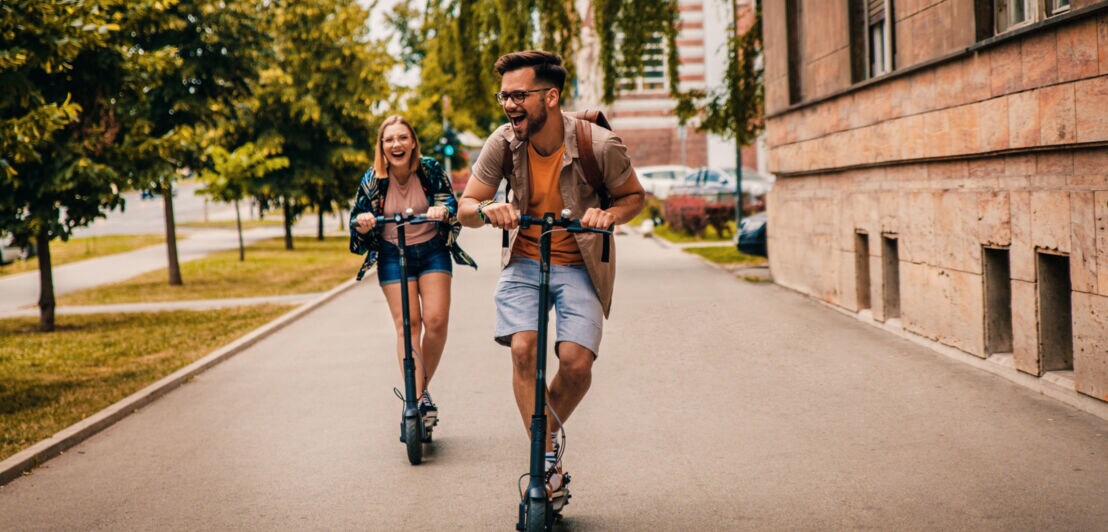 Ein junges Paar fährt lachend auf E-Scootern über einen breiten Gehweg