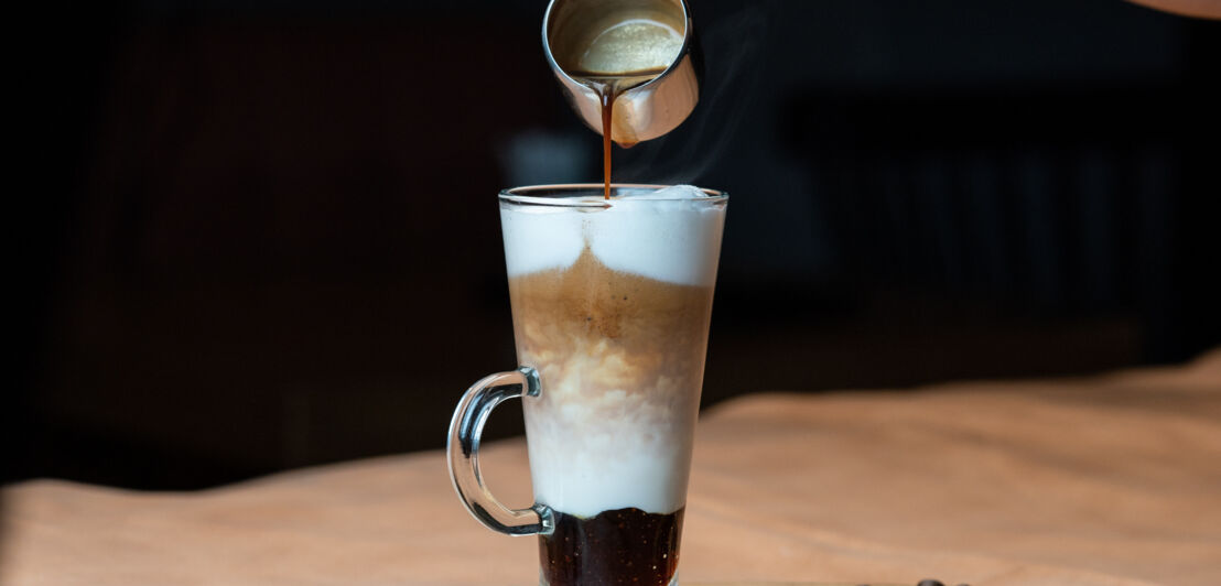 Espresso wird aus einem Kännchen in ein Glas mit Milchschaum gegossen