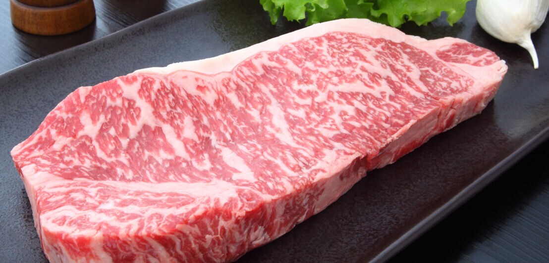Rohes Stück Fleisch vom Kobe-Rind auf einem schwarzen Teller mit einem Salatblatt und Knoblauch