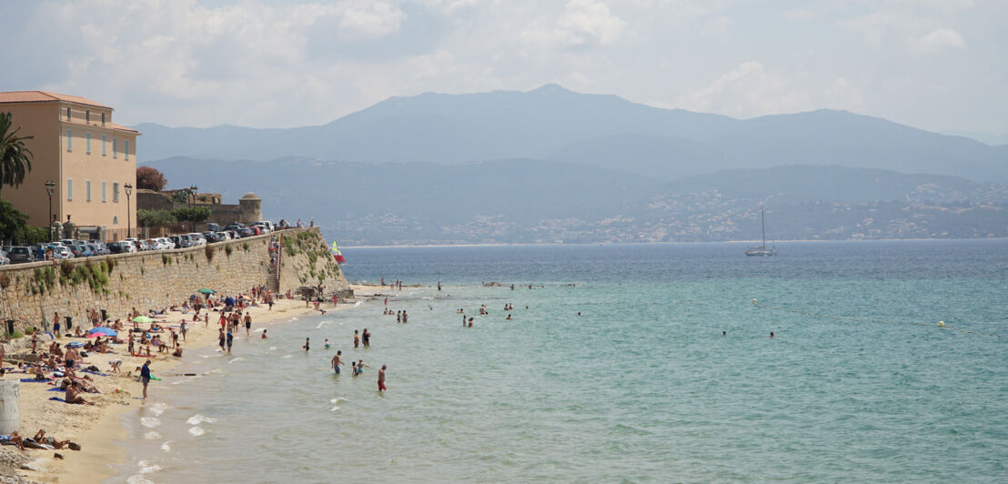 Ein Strandabschnitt mit badenden Menschen, im Hintergrund bergige Landschaft