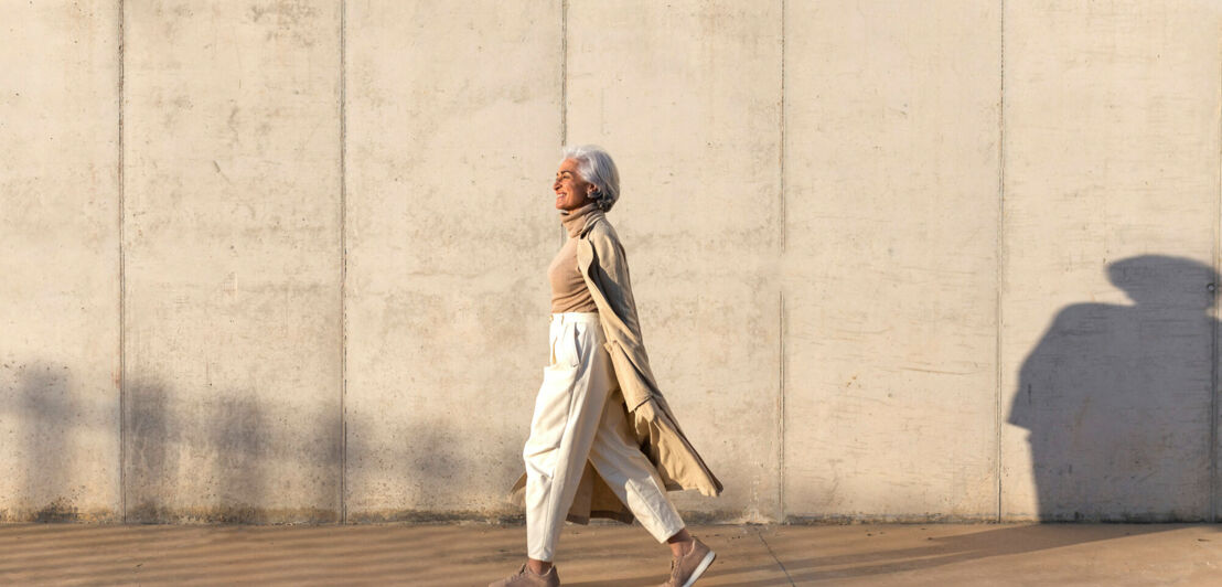 Eine ältere Frau in beiger Kleidung und langem Trenchcoat läuft vor einer Betonwand 