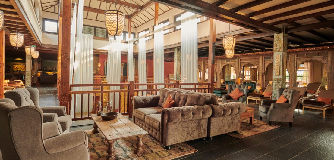 Der Innenbereich im vabali spa mit grauen Sitzgelegenheiten, weiß-transparenten Vorhängen, bunten Teppichen und einem Holzgeländer. 