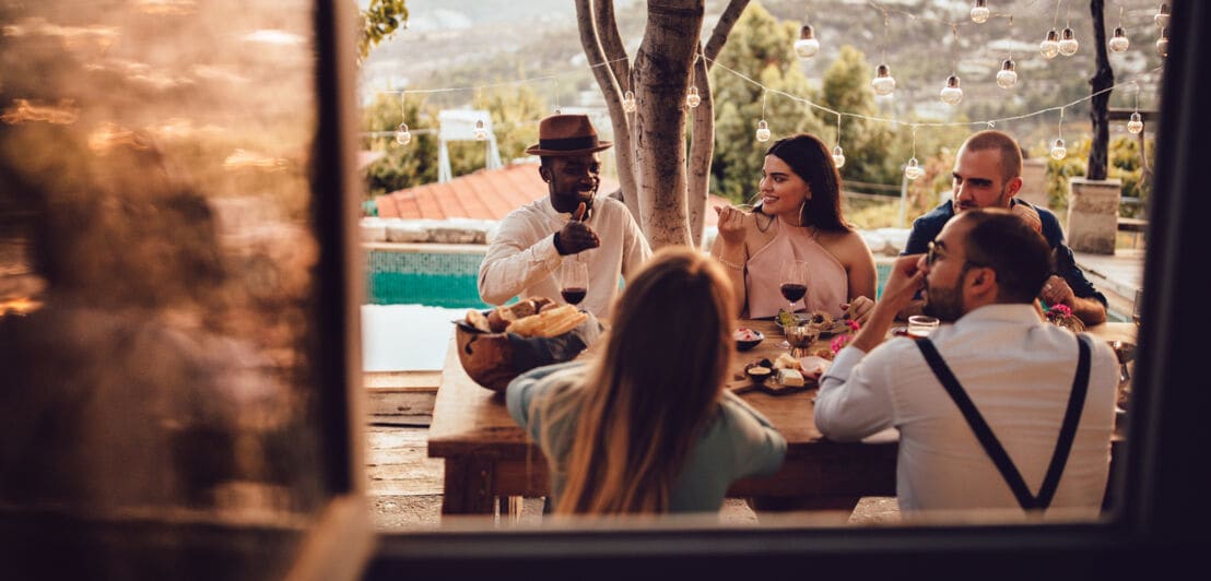 Eine Gruppe von Freunden genießt ein gemeinsames Dinner in zwangloser Atmosphäre an einem Tisch auf einer Terrasse mit Pool 