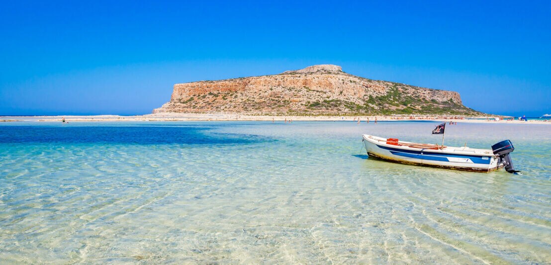 Lagunenstrand von Balos mit Blick auf türkisblaues Wasser, Motorboot und kleiner Insel am Horizont.