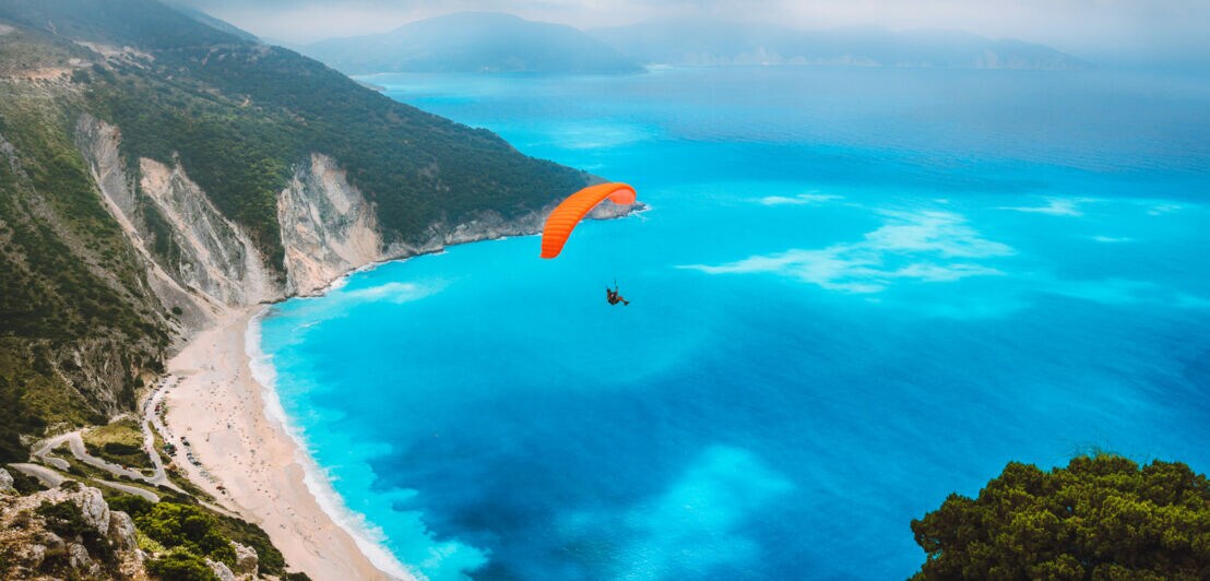 Ein Gleitschirmflieger über einer Badebucht an einer Bergküste mit Sandstrand und türkisblauem Wasser.