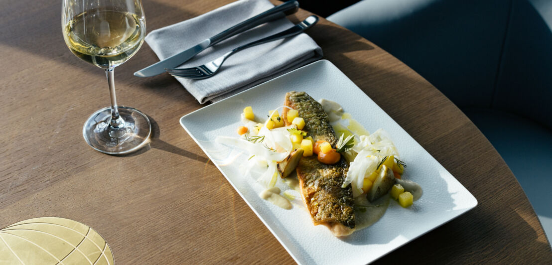 Ein Fischgericht auf einem rechteckigen Teller serviert steht auf einem Holztisch, dazu ein Glas Weißwein