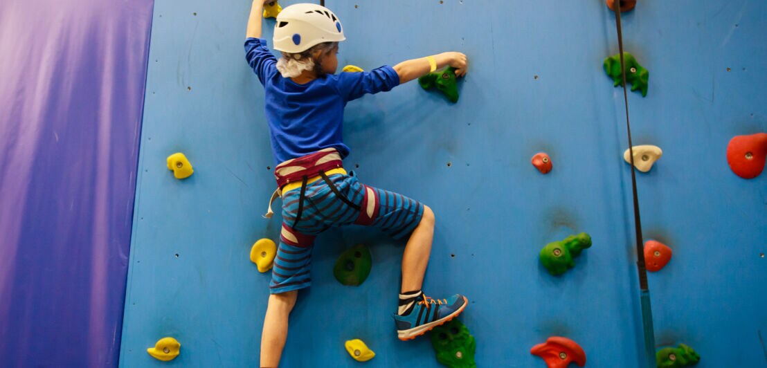 Ein kleiner Junge klettert mit einem Seil gesichert an einer Kletterwand hinauf