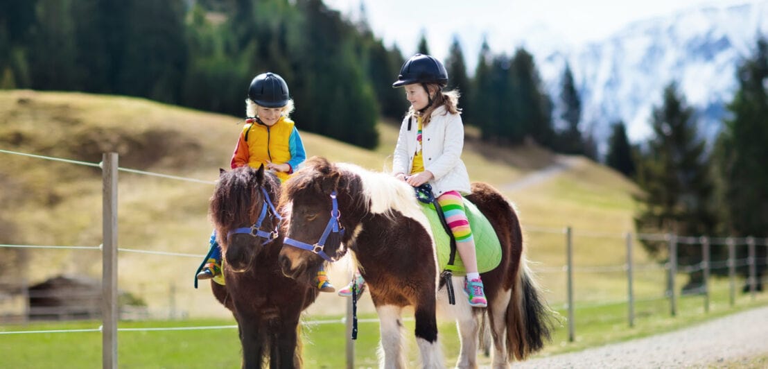 Zwei Kinder auf Ponys in ländlicher Umgebung vor Bergpanorama