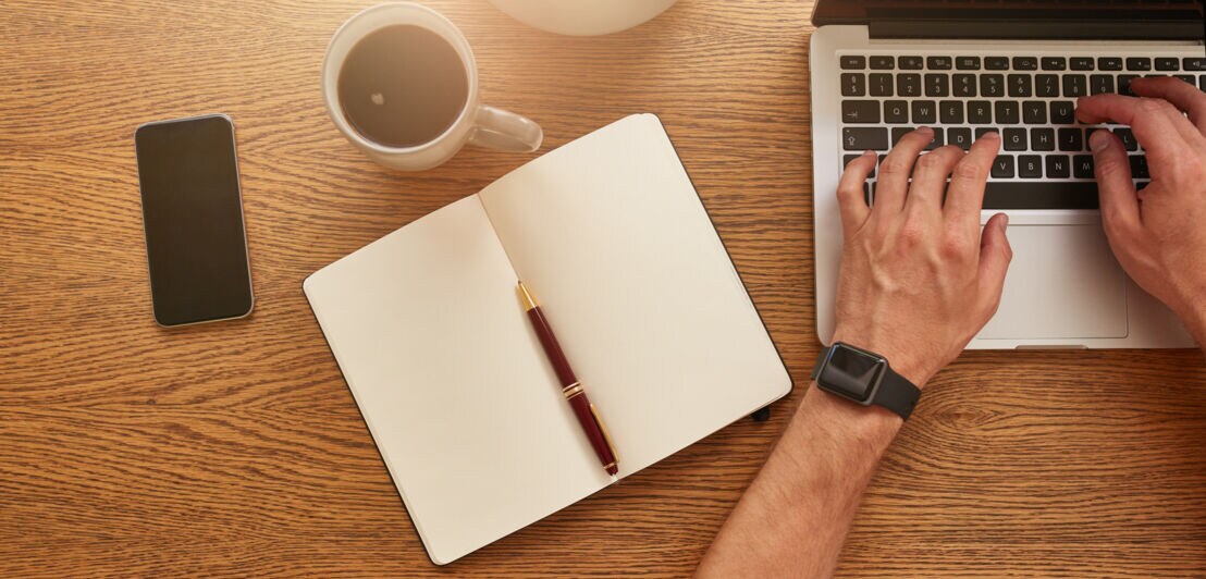 Sicht von oben auf einen Schreibtisch mit Kaffeetasse, Smartphone, Notizbuch und Kugelschreiber, ein Mann arbeitet an einem Notebook