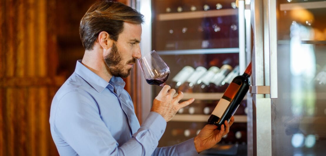 Ein Mann hält eine Rotweinflasche in der Hand und in der anderen ein Rotweinglas, an dem er riecht