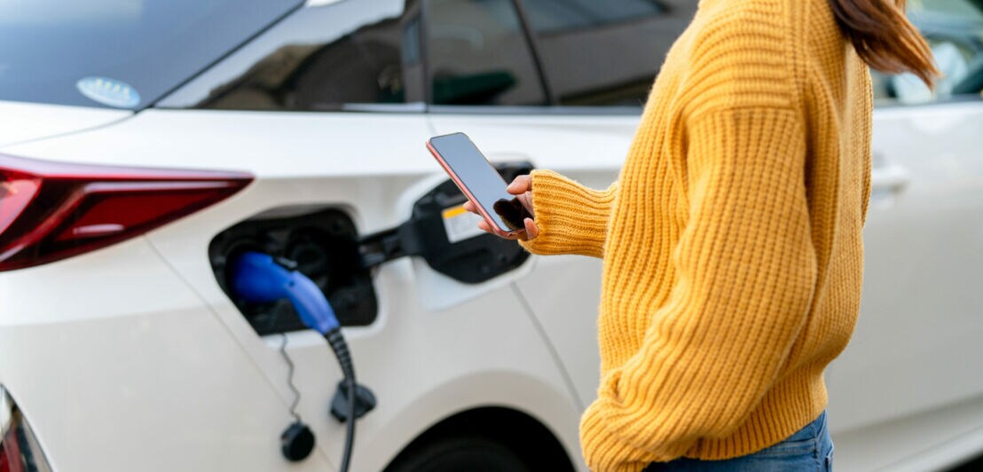 Eine Person hält ein Smartphone vor ein elektrisches Auto beim Ladevorgang