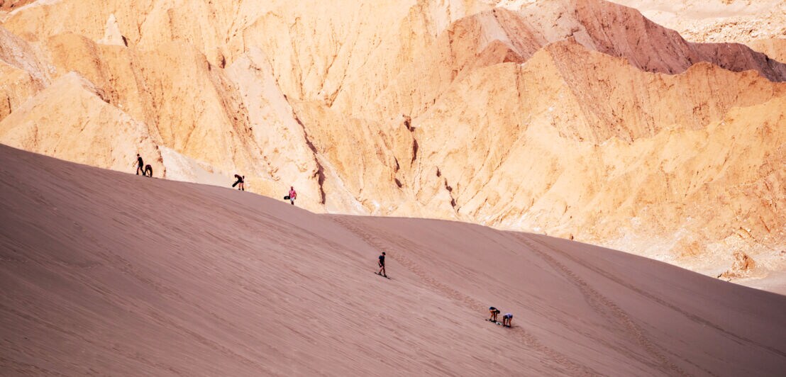 Personen auf Sandboards fahren eine Düne vor einer Felswand in einer Wüstenlandschaft hinunter
