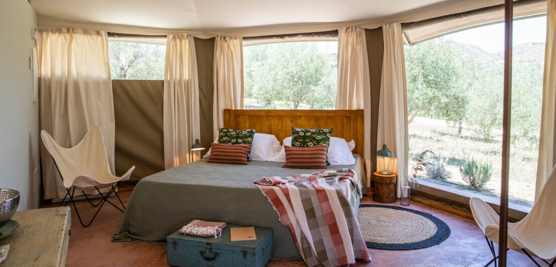 Bett in einer hellen Zelt-Lodge mit großen Fenstern, durch die Bäume und Hügel zu sehen sind
