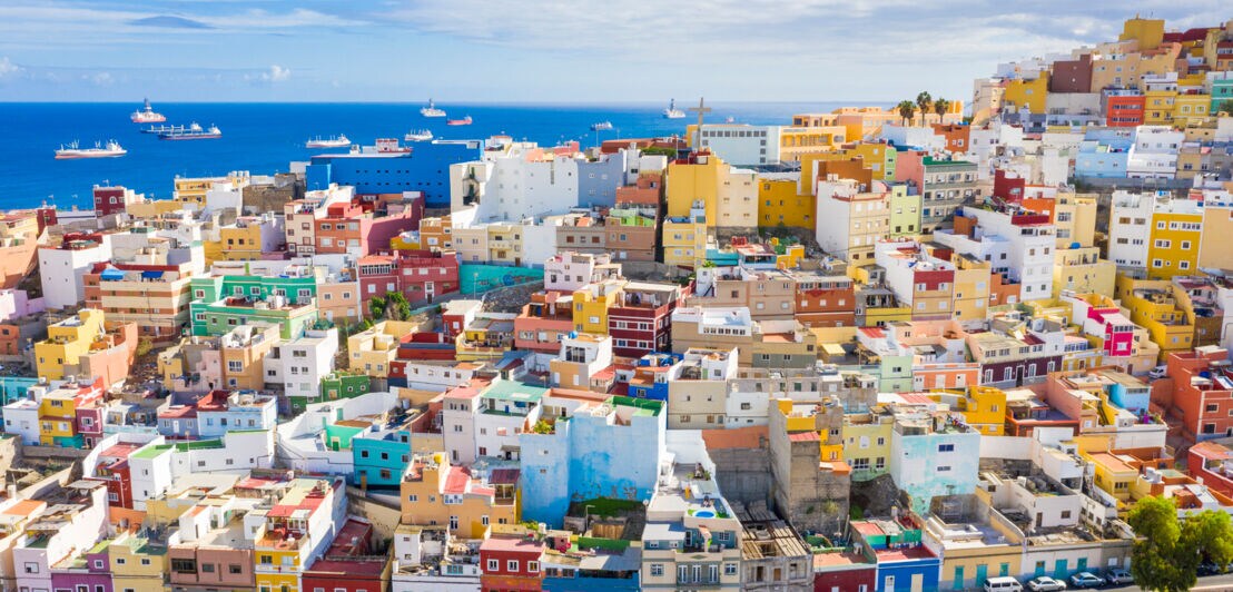 Stadtpanorama von Las Palmas mit bunten Häusern auf einem Hügel, im Hintergrund das Meer mit Schiffen
