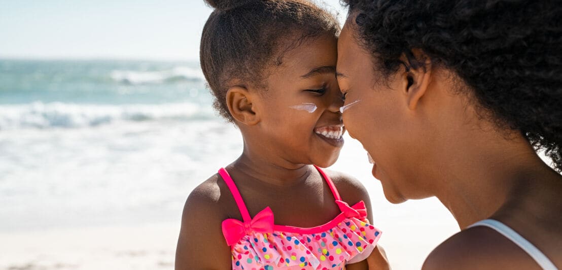 Ein kleines Mädchen am Strand mit Sonnencreme im Gesicht wird lachend von seiner Mutter umarmt