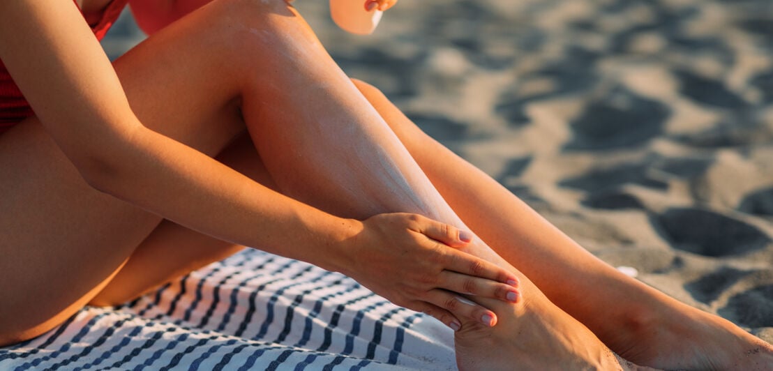 Eine junge Frau cremt ihre Beine mit Sonnencreme am Strand ein