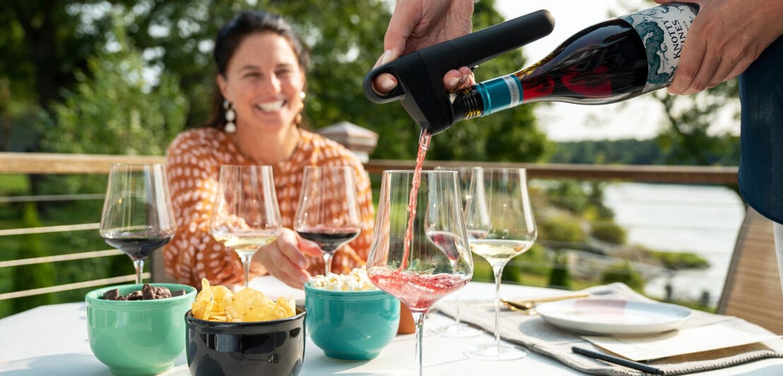 Eine Person gießt Rotwein aus einer Weinflasche mit einem Aufsatz in ein Glas auf einem gedeckten Tisch auf einer Terrasse