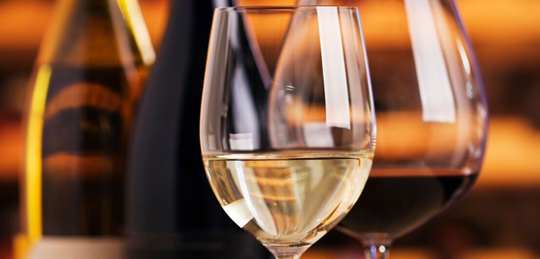 Nahaufnahme eines befüllten Rotweinglases und eines befüllten Weißweinglases vor zwei Weinflaschen