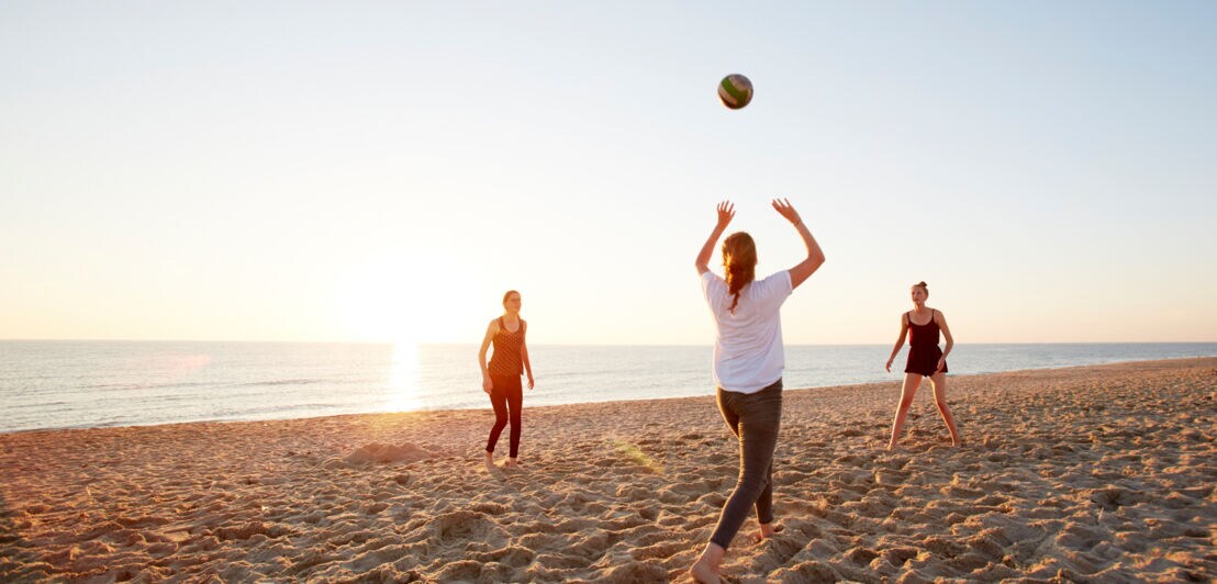 Drei Jugendliche spielen Volleyball an einem weiten Sandstrand bei Sonnenuntergang