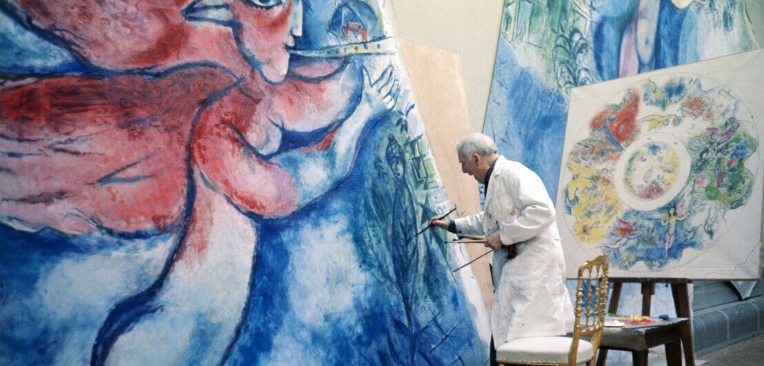 Marc Chagall malt in einem weißen Kittel an einer großen Leinwand, daneben eine Entwurfsskizze für ein Deckengemälde