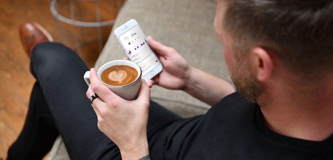 Ein Mann mit dunklem NFC-Ring hält einen Kaffee und ein Smartphone in der Hand.