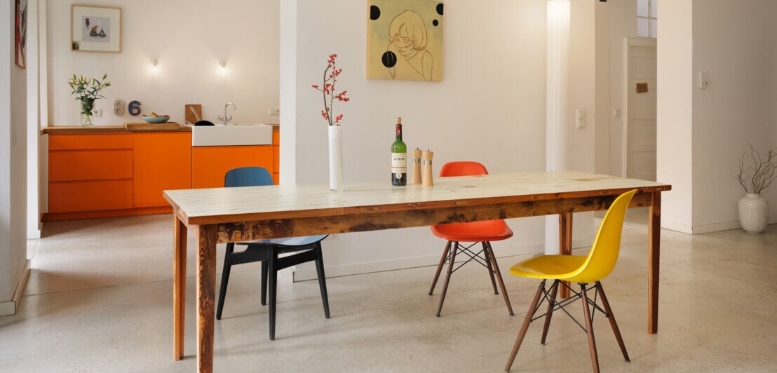 Blick in eine minimalistisch eingerichtete Wohnung mit Holztisch und drei bunten Stühlen.