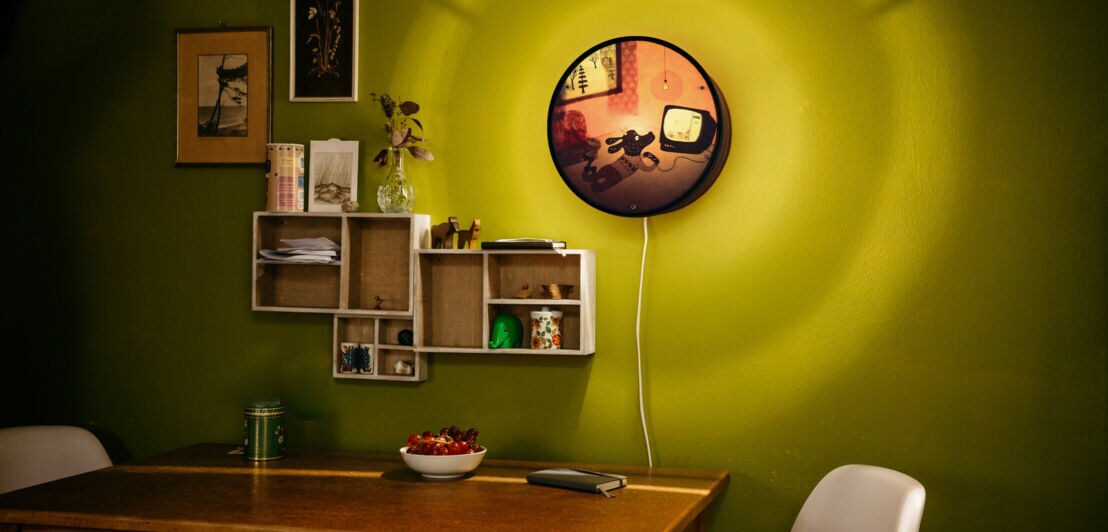 Ausschnitt einer Küche mit grüner Wand, Tisch, zwei Stühlen, ein paar kleinen Regalen an der Wand und einer Wandlampe.