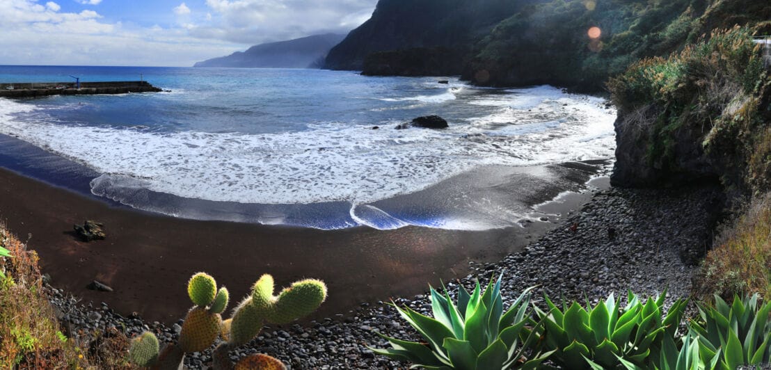 Panorama eines menschenleeren Strandes mit dunklem Sand und Steinen, umgeben von Felsen und tropischer Vegetation