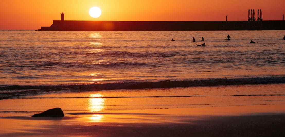 Personen treiben auf Surfbrettern am Strand im Wasser bei Sonnenuntergang