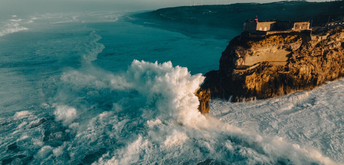 Luftaufnahme eines Küstenabschnitts mit Felsvorsprung, an dem hohe Wellen brechen