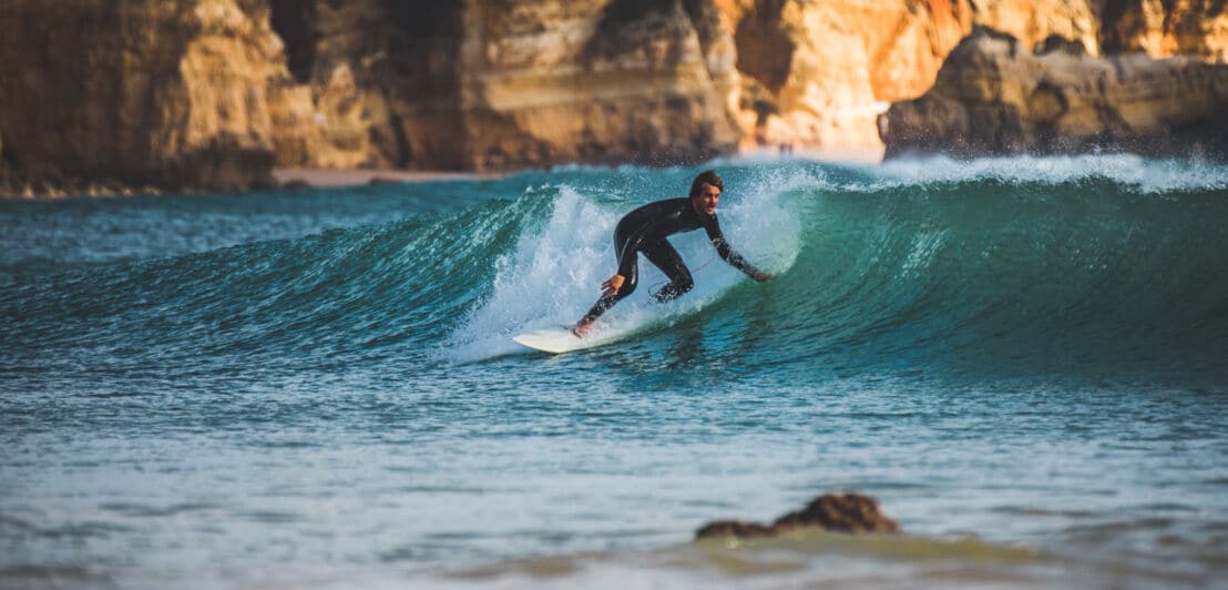 Ein Surfer im Neoprenanzug surft auf einer Welle im Meer vor einer Felsenküste