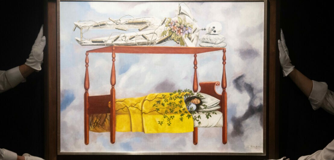 Vier Hände in weißen Handschuhen halten vorsichtig vor dunklem Hintergrund Frida Kahlos Bild „El sueño (La cama)“.