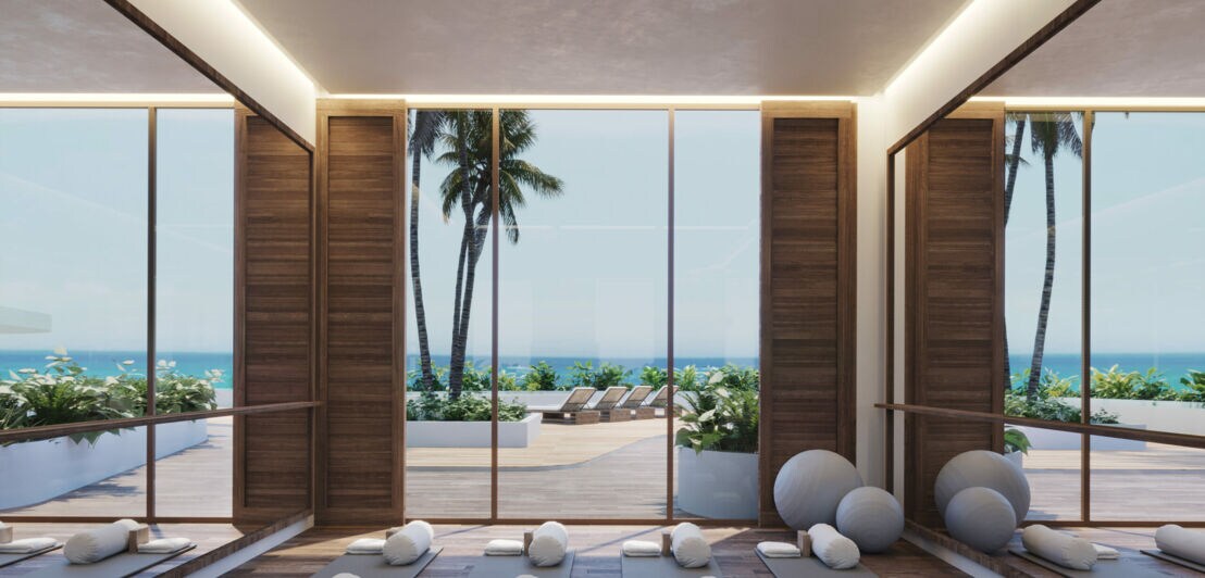 Ein Yogaraum mit offenen Fenstern vor denen Wasser und Palmen liegen
