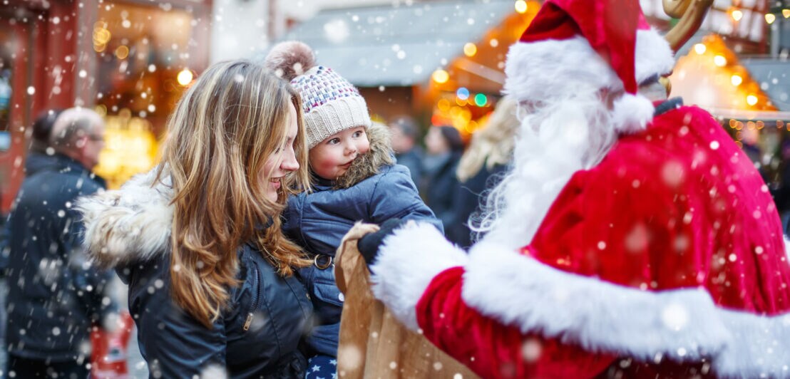 Ein Weihnachtsmann spricht zu einem kleinen Kind auf dem Arm seiner Mutter auf einem Weihnachtsmarkt