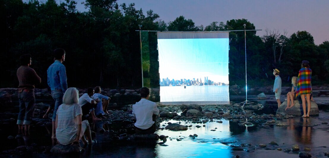 Mehrere Personen sitzen auf Steinen in einem Fluss und schauen auf eine kleine Kinoleinwand im Wasser.