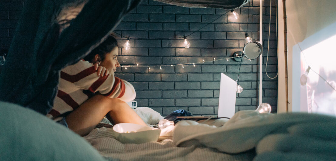 Eine junge Frau schaut im Bett einen Film, der von einem kleinen Beamer an eine Wand projiziert wird