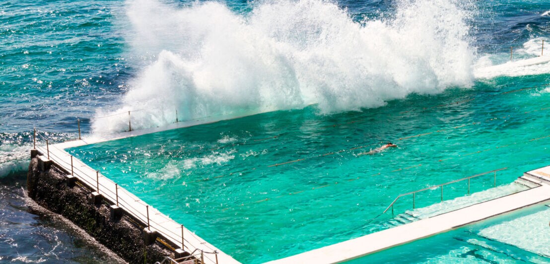 Männliche Athleten schwimmen in einem Meerwasserpool, an dem sich von außen die Wellen brechen.