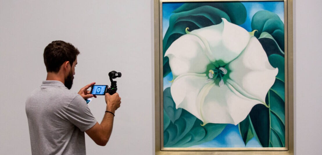 Ein Mann fotografiert ein Gemälde mit weißer Blüte der Künstlerin Georgia O‘Keeffe mit seinem Smartphone in einer Ausstellung