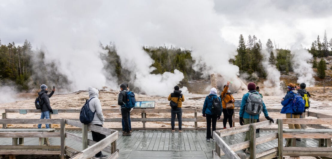 Menschen beobachten im Yellowstone-Nationalpark von einem Holzsteg aus dampfende Thermalquellen.