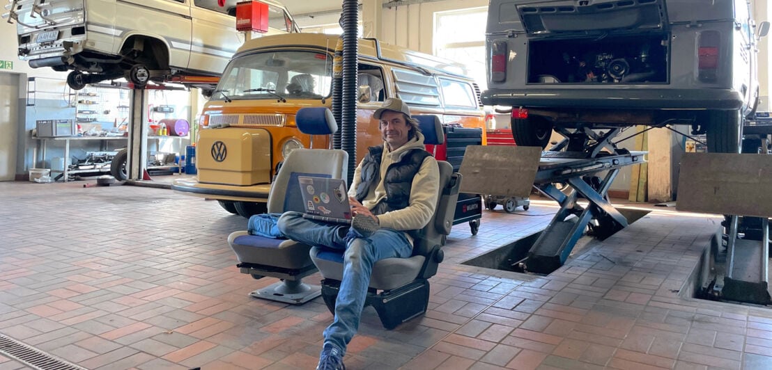 Ein Mann mit Laptop sitzt auf einem ausgebauten Fahrersitz in einer Autowerkstatt