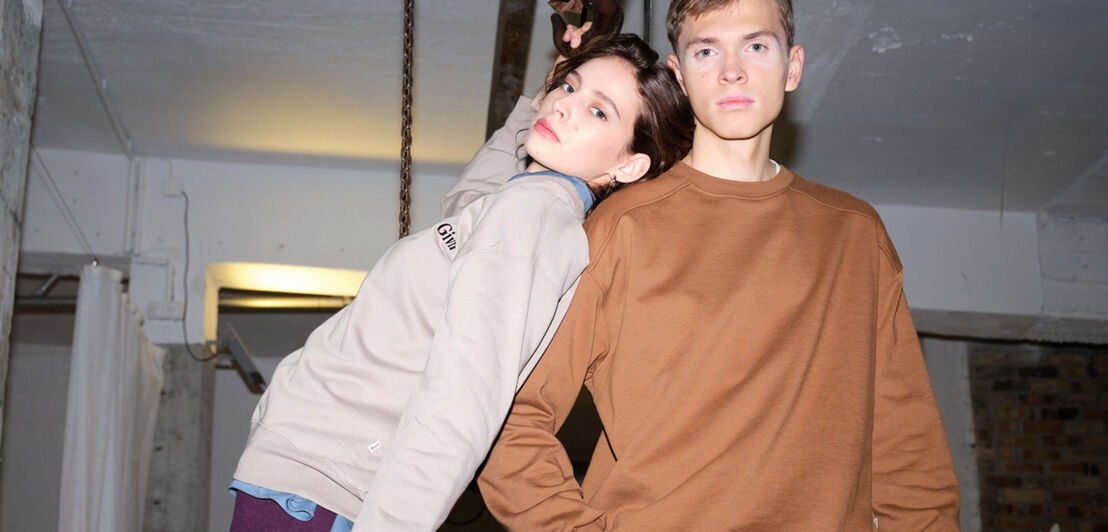 Zwei Models tragen schlichte Sweatshirts von Givn in Hellbeige und Rostbraun. Im Hintergrund ein industriell eingerichteter Raum.