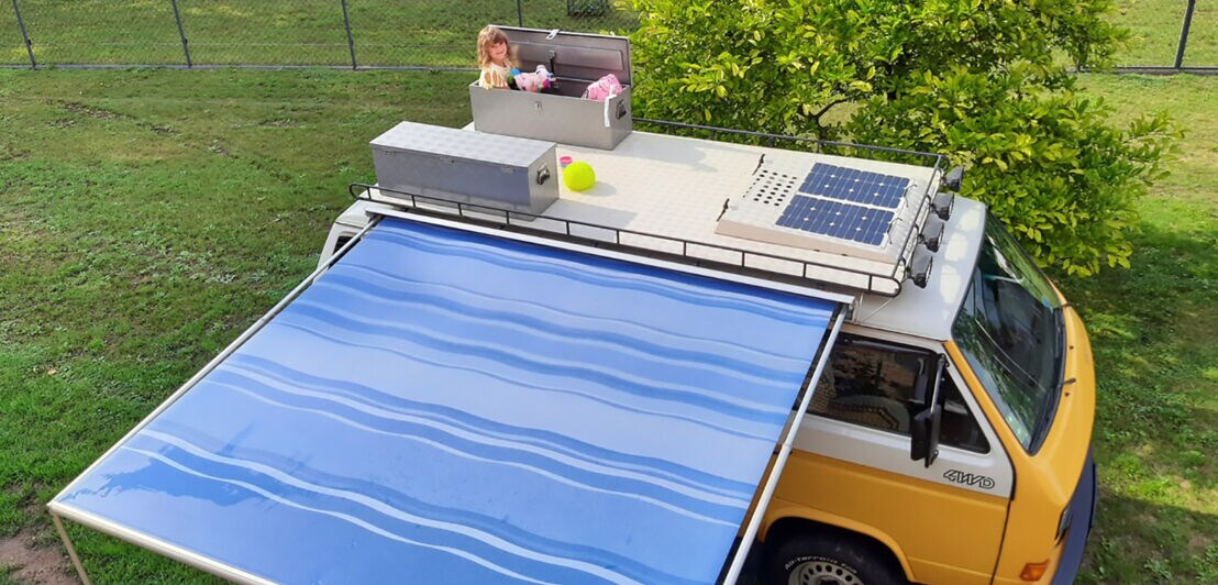 Blick von oben auf einen VW Bus mit Markise und einem Kind in einer Dachbox 