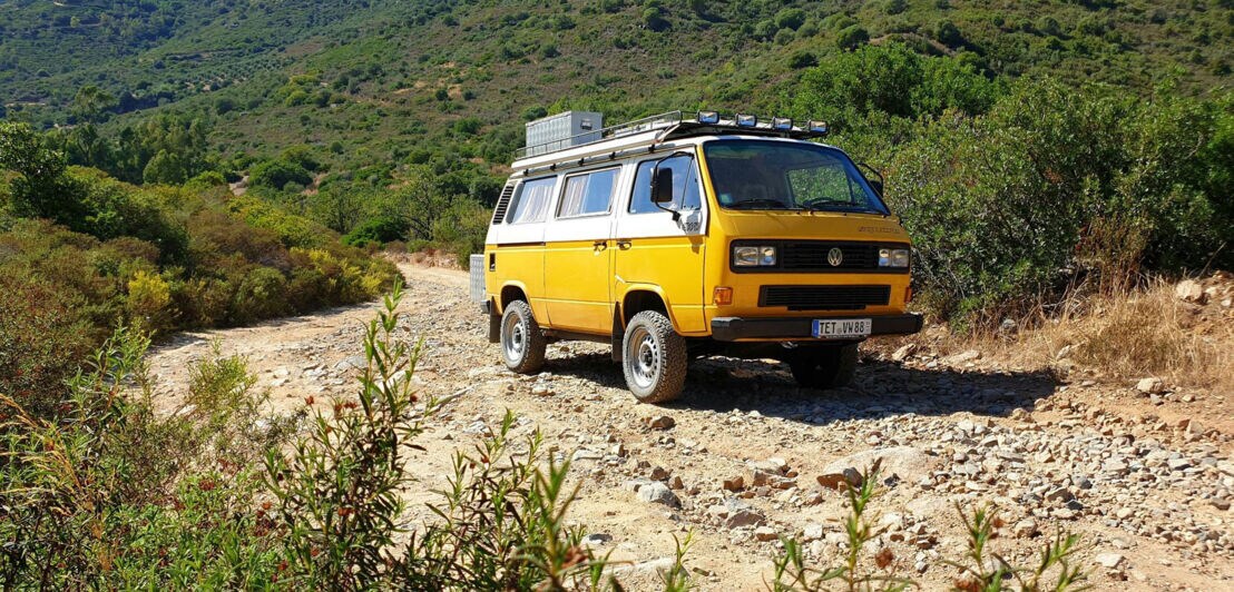 Ein gelber VW Bus auf einer Schotterstraße in einer bergigen Landschaft 