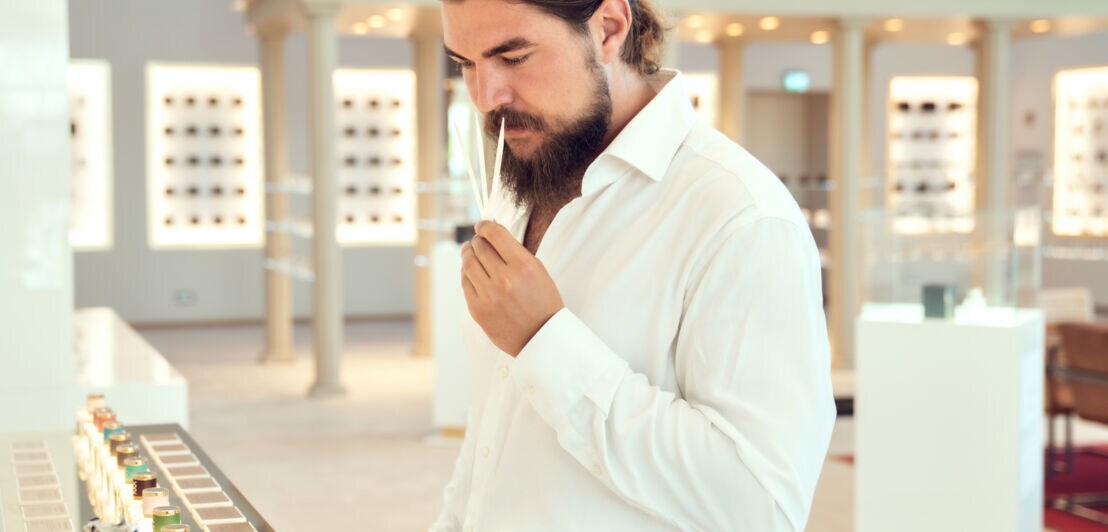 Ein Mann mit Vollbart in einem weißen Hemd riecht an drei Duft-Teststreifen im Showroom einer Parfümmanufaktur