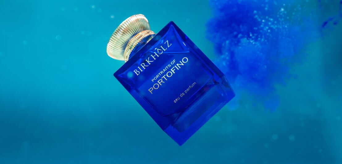 Ein eleganter, quadratischer Parfumflakon aus blauem Glas der Marke Birkholz mit goldenem Verschluss schwebt in blauem Wasser
