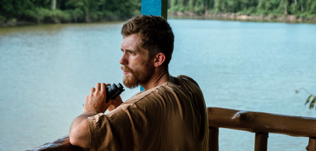 Ein junger Mann mit Bart steht an der Reling eines Hausbootes auf einem Fluss und schaut in den Regenwald, in der Hand hält er ein Fernglas.